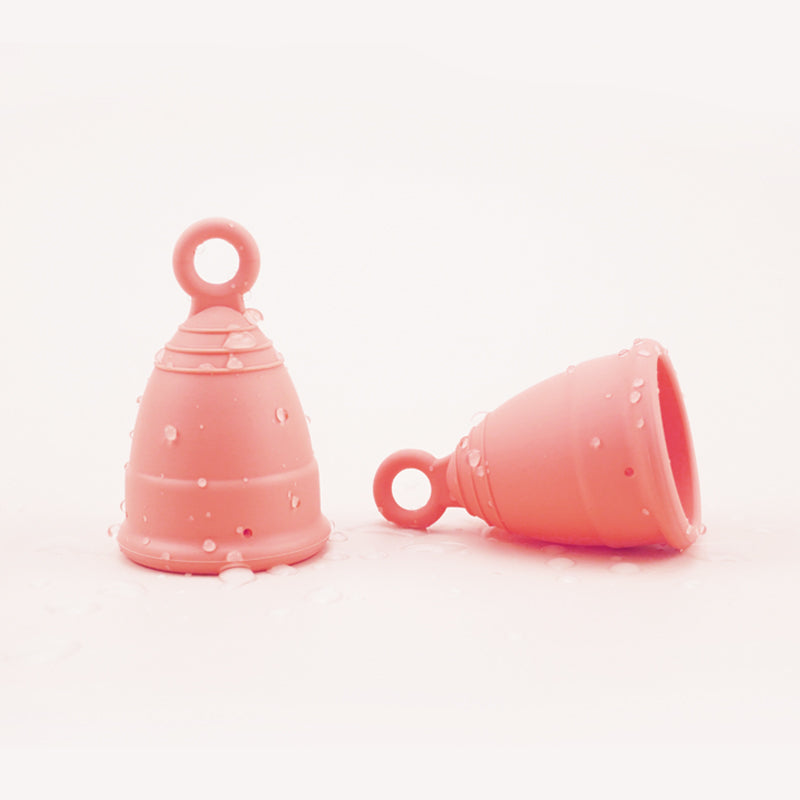 Two menstrual cups shown with water droplets in colour peach, one cup standing horizontally, one cup on its side, both cups have rings